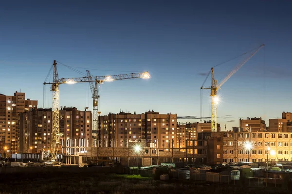building site with three tower cranes against the background of multi-storey houses and a dark blue sky, night scene