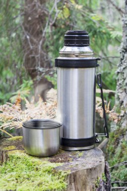 Çelik termos ve bir fincan sıcak tişört kozalaklı ormandaki eski kütüğün üzerinde duruyor, soyut arka planda kamp kuruyorlar.