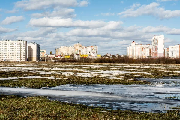 View of the field with a frozen pond and the outskirts of the city, new city under construction, spring sunny day — Stock Photo, Image
