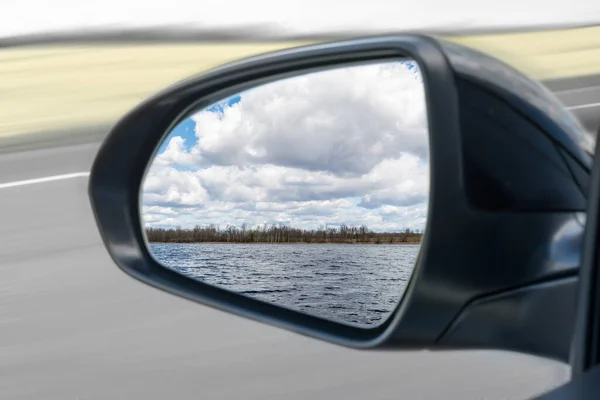 in the mirror of the car you can see a forest lake and large white clouds in autumn or spring