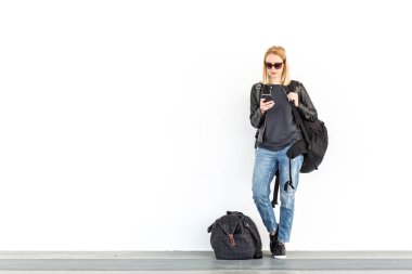Cep telefonu kullanırken, seyahat çantası yanında duran ve düz beyaz duvara İstasyonu whit üzerinde bekleyen moda genç kadın.