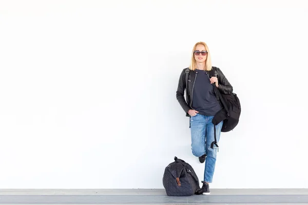 Mujer joven de moda de pie y esperando contra la pared blanca llanura en la estación whit bolsa de viaje a su lado . —  Fotos de Stock