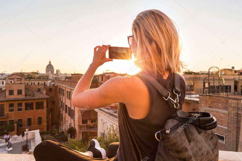 Female tourist taking mobile phone photo of Piazza di Spagna, landmark square with Spanish steps in Rome, Italy at sunset.