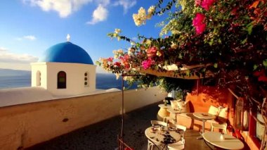 Tipik Yunan köyü Oia, Santorini Island, Yunanistan'ın geleneksel kafeteryada bir teras ile renkli çiçek Funda oynamak rüzgar ile mavi kilise kubbe.