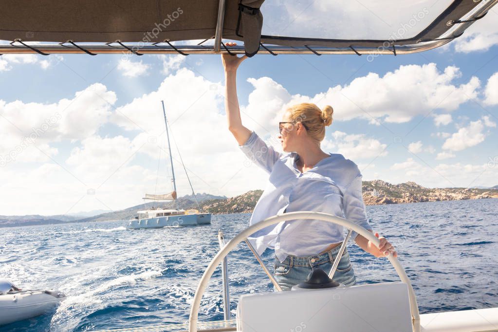 Attractive blond female skipper navigating the fancy catamaran sailboat on sunny summer day on calm blue sea water.