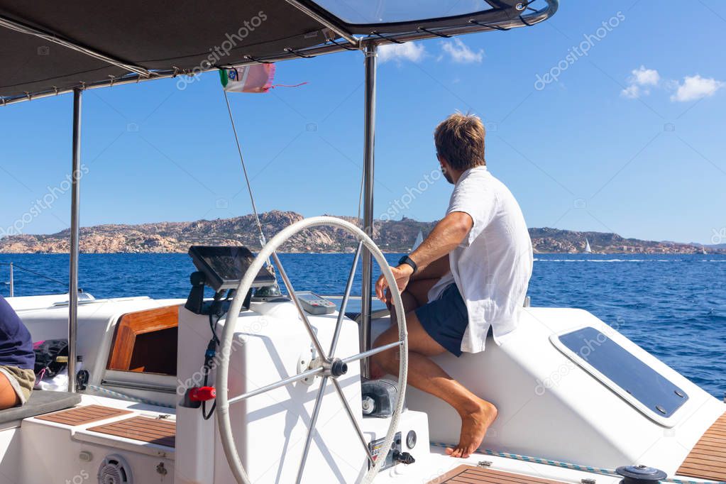 Male skipper navigating the fancy catamaran sailboat on sunny summer day on calm blue sea water.