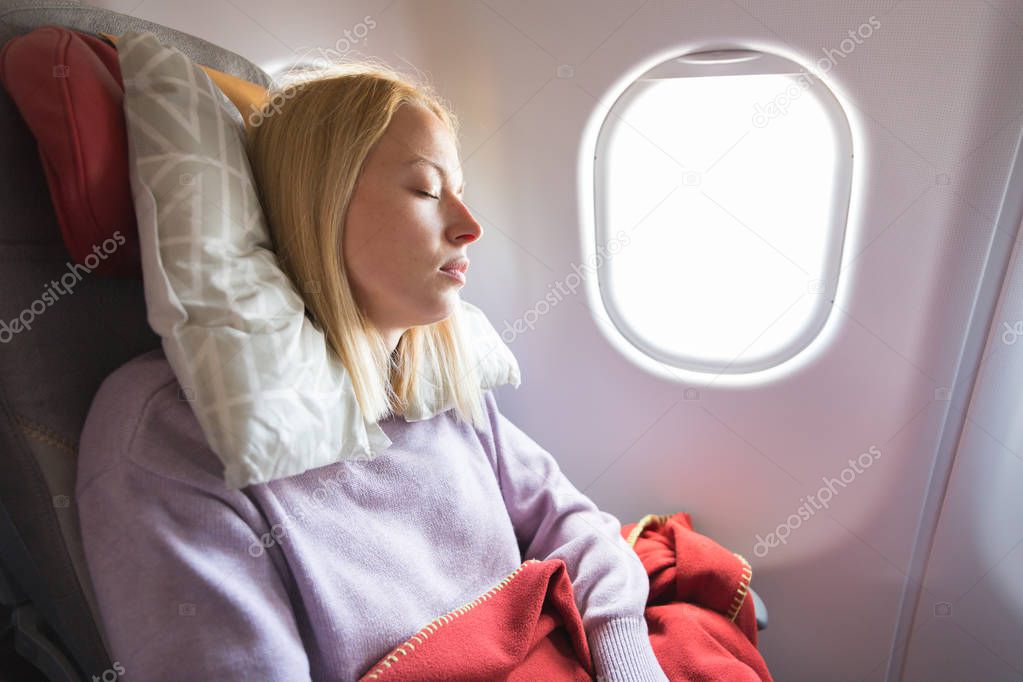 Tired blonde casual caucasian lady napping on seat while traveling by airplane. Commercial transportation by planes.
