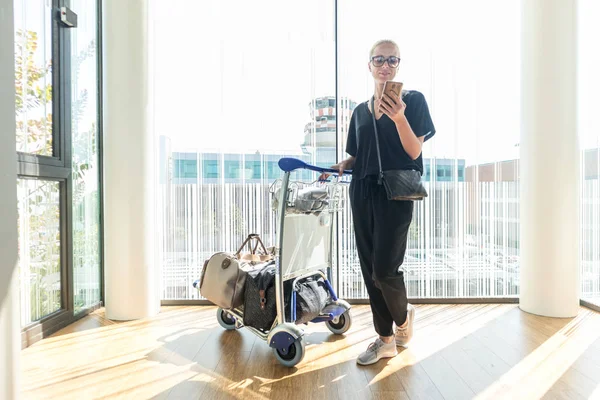 Casual kvinna på flygplatsterminalen talar på mobilen, väntar på avgång med bagage på vagnens varukorg. — Stockfoto