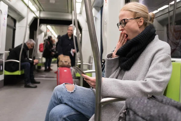 Hermosa mujer rubia bostezando mientras lee por teléfono, viajando en metro. Transporte público . —  Fotos de Stock