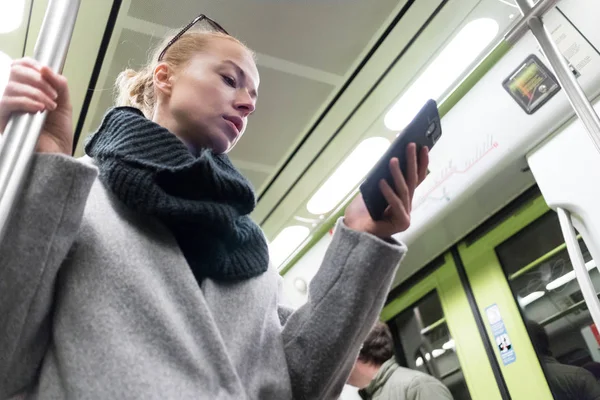 Schöne blonde Frau in Wintermantel und Schal, die am Telefon liest, während sie mit öffentlichen Verkehrsmitteln unterwegs ist. — Stockfoto