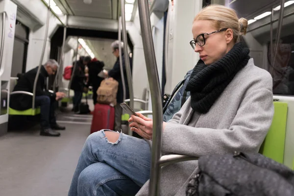 Hermosa mujer rubia con abrigo de invierno y bufanda leyendo por teléfono mientras viaja en transporte público de metro . — Foto de Stock