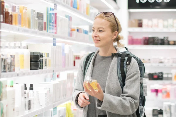 Blond young female traveler wearing travel backpack choosing perfume in airport duty free store.