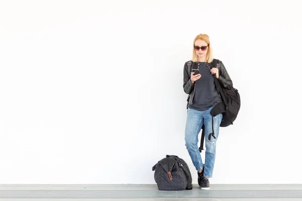 Divatos fiatal nő, a mobiltelefon, míg állandó, és az állomás Pünkösd sima fehér falhoz vár utazik táska oldalán. — Stock Fotó
