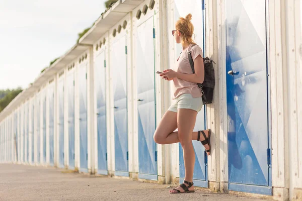 Blonde young female traveler wearing summer style clothing, holding mobile phone, against retro blue beach dressing rooms at summer time vacation. — Stock Photo, Image
