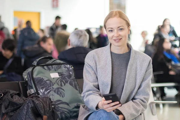 Веселая женщина-путешественница улыбается, смотрит в камеру, читая по мобильному телефону, ожидая посадки в самолет на выходе из терминала аэропорта . — стоковое фото