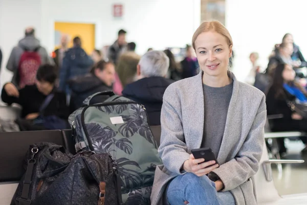 Vidám női utas mosolyogva, látszó-on fényképezőgép olvasásakor a mobilján, miközben arra vár, hogy fedélzetén a repülőgép indulási kapuk reptéren. — Stock Fotó