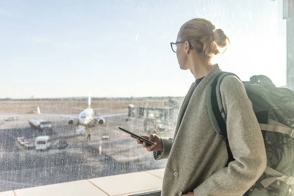 Havaalanında sıradan giyinmiş kadın gezgin, elinde akıllı telefon cihazıyla havaalanı pistindeki uçakların kapı camlarından içeri bakıyor. — Stok fotoğraf