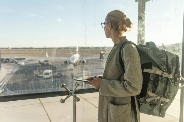 Satunnaisesti pukeutunut naispuolinen matkustaja lentokentällä, jolla on älypuhelin laite, katselee lentokentän portin ikkunoiden läpi lentokenttien kiitoradalla — kuvapankkivalokuva
