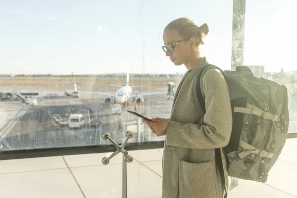 Viaggiatore femminile vestito con disinvoltura in aeroporto guardando il dispositivo smart phone di fronte alle finestre del cancello dell'aeroporto con vista aerei sulla pista dell'aeroporto — Foto Stock