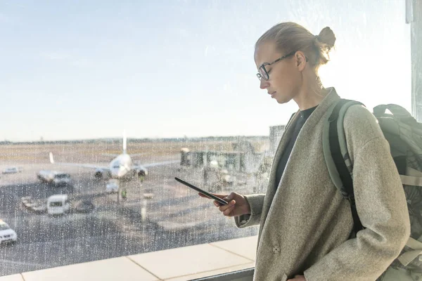 Viajera casualmente vestida en el aeropuerto mirando el dispositivo de teléfono inteligente frente a las ventanas de la puerta del aeropuerto con vistas a los aviones en la pista del aeropuerto —  Fotos de Stock
