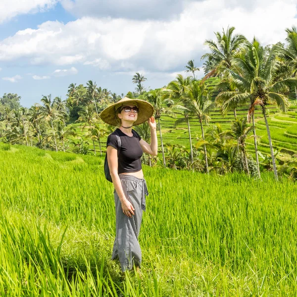 Расслабленная модная кавказская туристка в маленьком рюкзаке и традиционной азиатской шляпе, гуляющая среди красивых зеленых рисовых полей и террас на острове Бали — стоковое фото