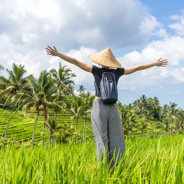Entspannte modische Reiserückkehrerin mit kleinem Rucksack und traditionellem asiatischen Reismütze, die Arme in den Himmel gereckt, genießt Natur pur auf wunderschönen grünen Reisfeldern und Terrassen auf der Insel Bali — Stockfoto