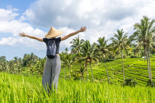 Entspannte modische Reiserückkehrerin mit kleinem Rucksack und traditionellem asiatischen Reismütze, die Arme in den Himmel gereckt, genießt Natur pur auf wunderschönen grünen Reisfeldern und Terrassen auf der Insel Bali — Stockfoto