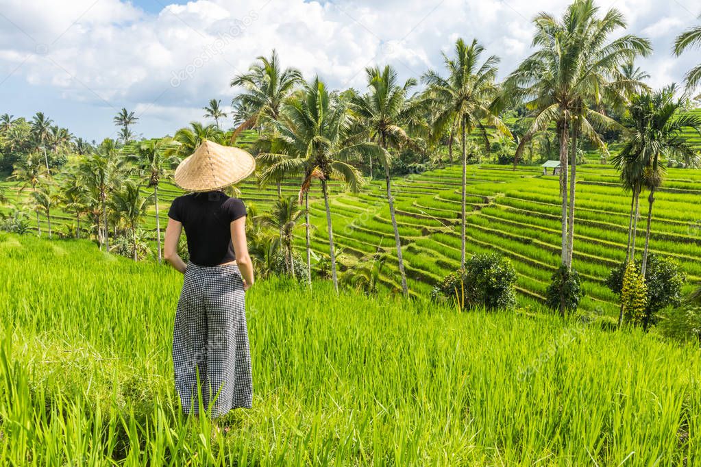 Relaxed fashionable caucasian female tourist wearing small backpack and traditional asian paddy hat looking at beautiful green rice fields and terraces on Bali island