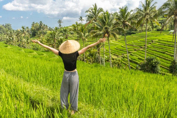 Entspannte modische Reiserückkehrerin mit kleinem Rucksack und traditionellem asiatischen Reismütze, die Arme in den Himmel gereckt, genießt Natur pur auf wunderschönen grünen Reisfeldern und Terrassen auf der Insel Bali — Stockfoto