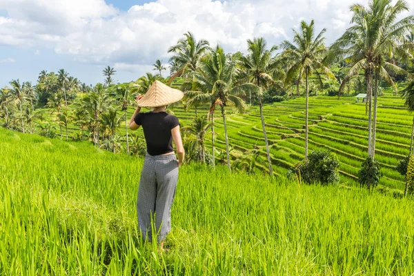 Расслабленная модная кавказская туристка в маленьком рюкзаке и традиционной азиатской шляпе с видом на красивые зеленые рисовые поля и террасы на острове Бали — стоковое фото