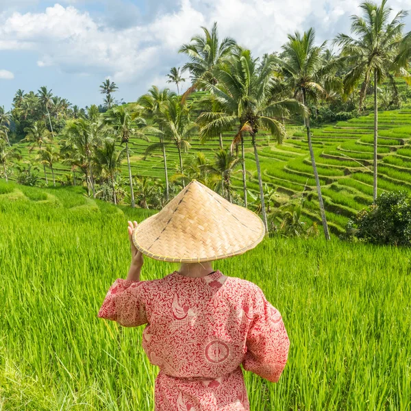 Relaxed fashionable caucasian woman wearing red asian style kimono and traditional asian paddy hat walking amoung beautiful green rice fields and terraces on Bali island