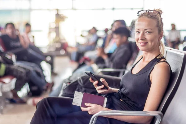 Casual tanned blond female traveler holding cell phone, passport and boarding pass while waiting to board a plane at the departure gates at the asian airport terminal — Stock Photo, Image