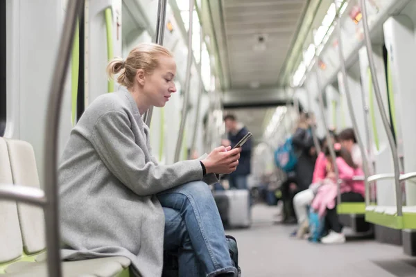 Hermosa mujer rubia con abrigo de invierno leyendo por teléfono mientras viaja en metro transporte público . — Foto de Stock