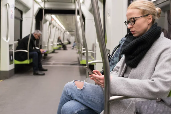 Hermosa mujer rubia con abrigo de invierno y bufanda leyendo por teléfono mientras viaja en transporte público de metro . — Foto de Stock