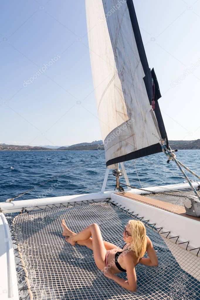 Beautiful woman relaxing on a summer sailing cruise, lying and sunbathing in hammock of luxury catamaran sailing around Maddalena Archipelago, Sardinia, Italy in warm afternoon light