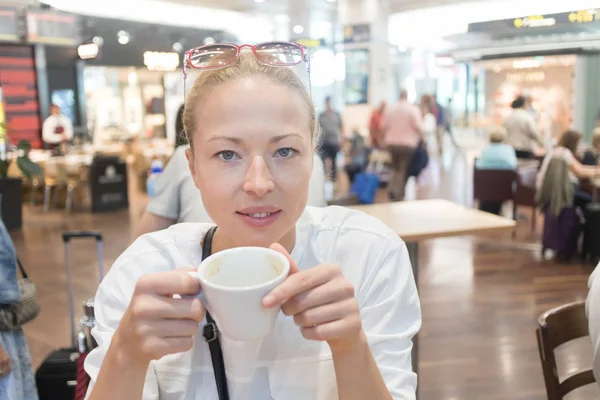 Πορτρέτο μιας περιστασιακής νεαρής ξανθιάς που έχει ένα φλιτζάνι καφέ, που κάθεται σε ένα καφέ στο εσωτερικό ενός αεροδρομίου, σταθμού, αγοράς τροφίμων ή εμπορικού κέντρου. — Φωτογραφία Αρχείου