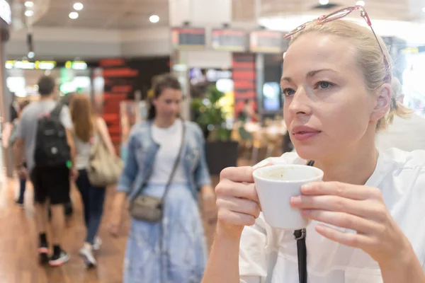 Retrato de uma jovem mulher loira casual tomando uma xícara de café, sentada no café dentro de um aeroporto, estação, mercado de alimentos ou shopping center . — Fotografia de Stock