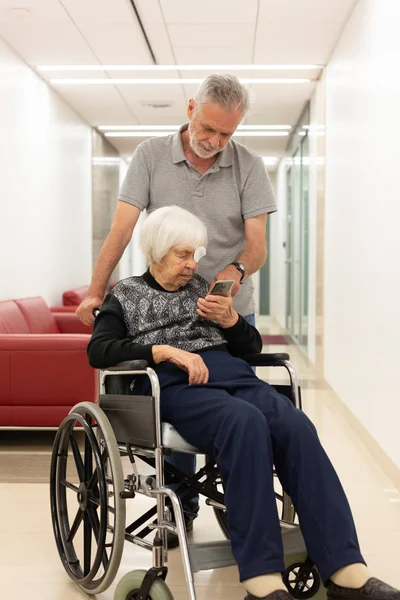 Středního věku člověka ukazuje a pomoc starších 95 let stará žena sedící na vozíku jak používat moderní mobilní telefon. — Stock fotografie