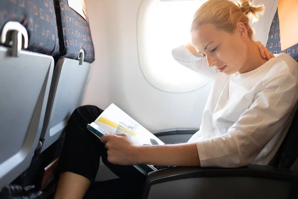 Mulher sentindo dor no pescoço ao ler na revista de voo em voo de avião intercontinental longo. Feminino viajante desconfortável sentado na cabine de passageiros . — Fotografia de Stock