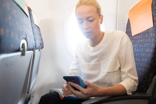 Молодая женщина использует мобильный телефон в самолете. Женщина-путешественница читает по телефону на сиденье самолета возле окна во время полета в самолете. Светит солнце через окно самолета — стоковое фото