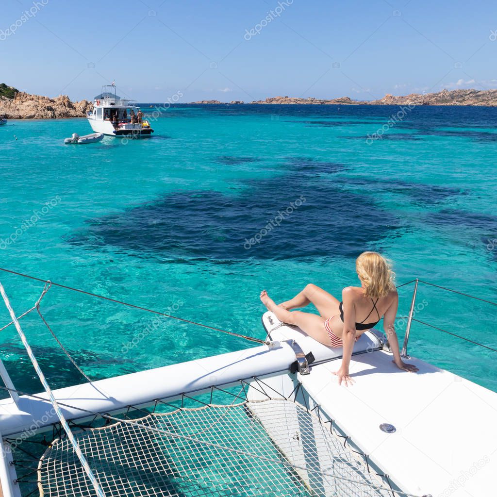 Woman relaxing on a summer sailing cruise, sitting on a luxury catamaran near picture perfect white sandy beach on Spargi island in Maddalena Archipelago, Sardinia, Italy.