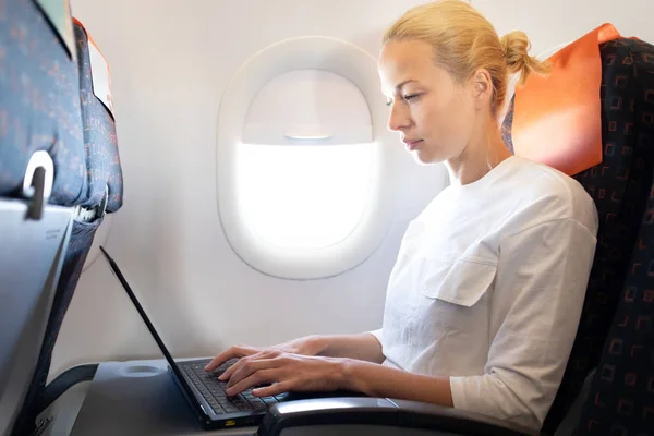 Atrakcyjny kaukaski żeński pasażer pracujący na nowoczesnym komputerze przenośnym przy użyciu połączenia bezprzewodowego na pokładzie samolotu komercyjnego lotu — Zdjęcie stockowe