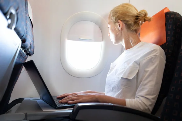 Attraktiv kaukasisk kvinnlig passagerare tittar genom fönstret medan du arbetar på modern bärbar dator med hjälp av trådlös anslutning ombord på kommersiella flygplan flygning — Stockfoto