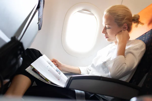 Woman reading in flight magazine on airplane. Female traveler reading seated in passanger cabin. Sun shining trough airplane window — 스톡 사진