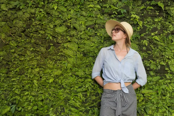 Retrato de una hermosa viajera. Mujer joven sonriente en sombrero de verano con gafas de sol, de pie frente a la exuberante vegetación tropical fondo de la pared . —  Fotos de Stock