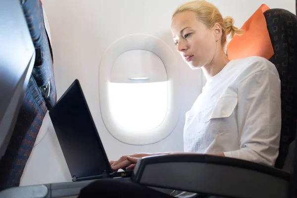 Привлекательная кавказка-пассажирка, работающая за современным ноутбуком с использованием беспроводной связи на борту коммерческого самолета — стоковое фото