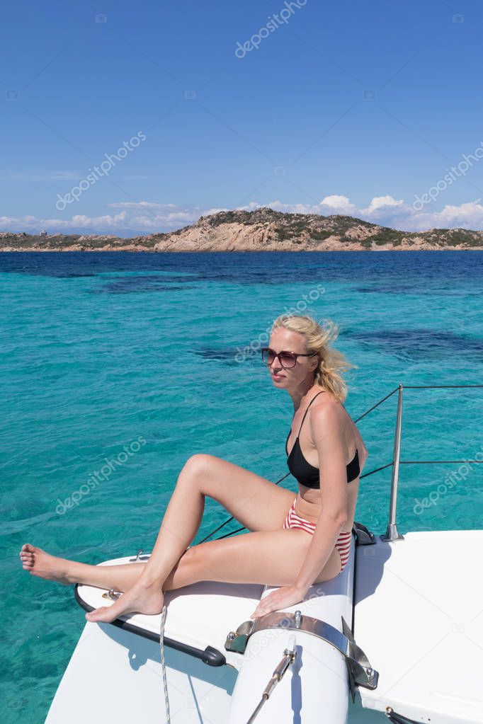 Woman relaxing on a summer sailing cruise, sitting on a luxury catamaran near picture perfect white sandy beach on Spargi island in Maddalena Archipelago, Sardinia, Italy.