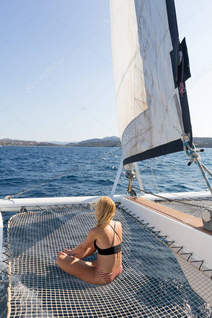 Beautiful woman relaxing on a summer sailing cruise, sitting and sunbathing in hammock of luxury catamaran sailing around Maddalena Archipelago, Sardinia, Italy in warm afternoon light