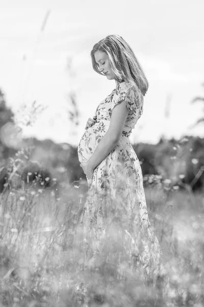 黄色の花の花でいっぱいの草原で白い夏のドレスで美しい妊娠中の女性. — ストック写真
