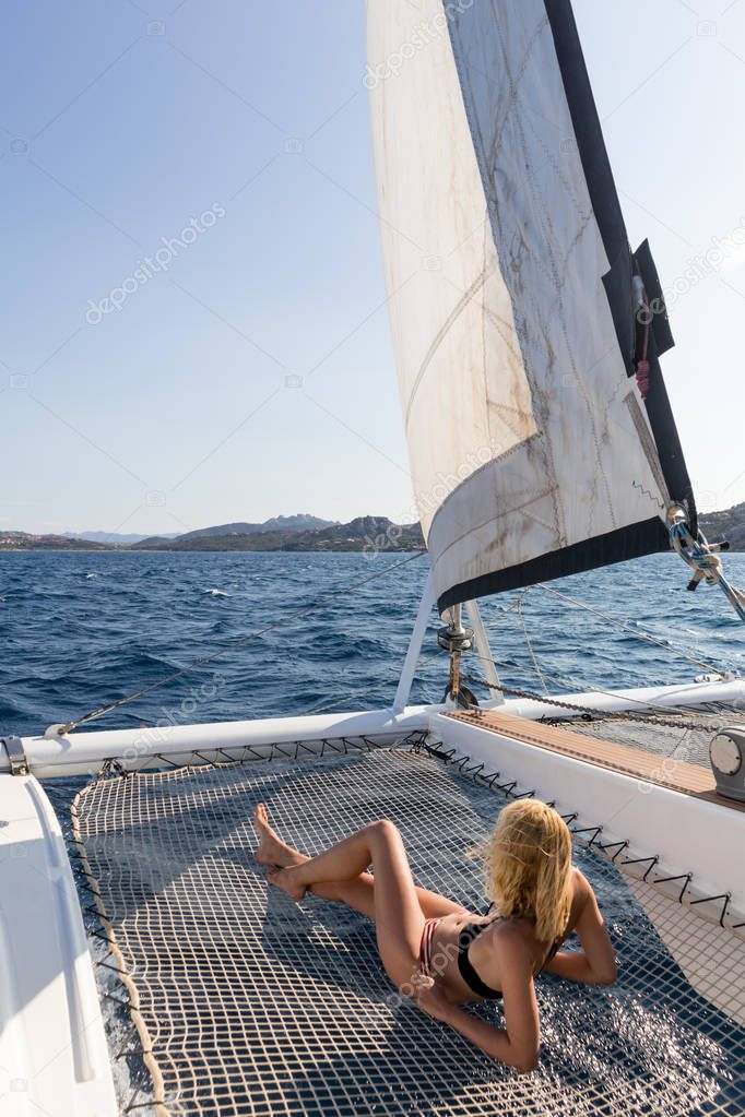 Beautiful woman relaxing on a summer sailing cruise, lying and sunbathing in hammock of luxury catamaran sailing around Maddalena Archipelago, Sardinia, Italy in warm afternoon light